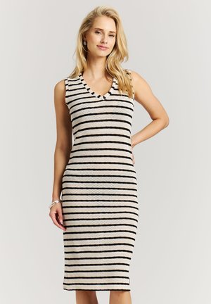 Blonde vrouw die een mouwloze, knielange, zwart-wit gestreepte gebreide jurk draagt, staand met één hand op haar heup.