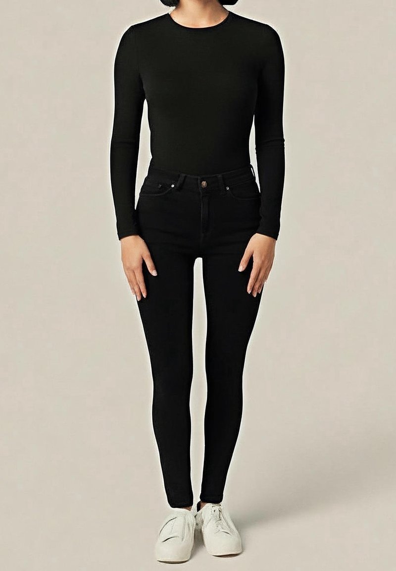Personne portant un haut noir à manches longues et un jean skinny noir avec des baskets blanches à lacets, debout devant un fond uni.