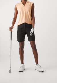 Koszulka polo bez rękawów z kołnierzykiem w kolorze brzoskwiniowym, czarne szorty, białe buty golfowe oraz szara rękawica golfowa, trzymająca kij golfowy na jednolitym tle.