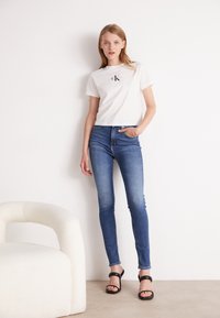 Bílá bavlněná tričko s logem "CK", v kombinaci s modrými skinny džíny. Modelka má na sobě černé sandály na pásky. Pozadí je jednoduché.