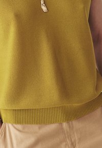 Gros plan sur un haut en tricot jaune moutarde avec un ourlet côtelé, porté avec un pantalon beige et un collier pendentif abstrait en or.