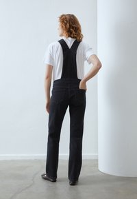 Svarta denimoveraller med rak passform, breda axelremmar och bakfickor, i kombination med en vit t-shirt. Slät yta, minimalistisk design.