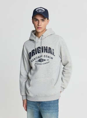 Giovane uomo che indossa una felpa con cappuccio grigio chiaro con la scritta "ORIGINAL Heritage Denim USA 1983" e un berretto blu navy con testo bianco abbinato.