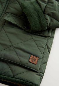 Grøn quiltet jakke med en kordfløjlskrave, brun læderpatch med logo og en glat tekstur. Designet har diamantformet syninger.