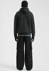 Schwarzer übergroßer Hoodie mit Kapuze, kombiniert mit lockeren schwarzen Cargo-Hosen mit Seitentaschen. Das Material scheint weich und langlebig zu sein.