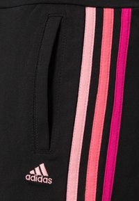 Černé sportovní kalhoty s boční kapsou a třemi vertikálními pruhy v odstínech růžové. Zahrnují vyšívané logo adidas v světle růžové barvě.