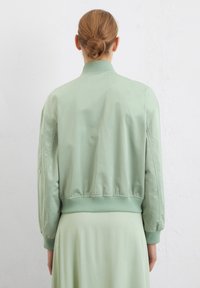 Giacca bomber verde chiaro con texture liscia, orlo e polsini a coste, e colletto classico. Nessuna ferramenta o motivo visibile.