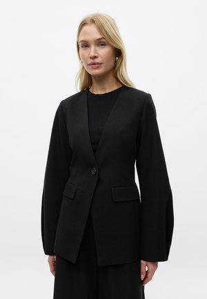 Lyshåret kvinde iført en skræddersyet sort blazer med en enkelt knap og klaplommer, stående mod en ensartet hvid baggrund.