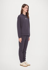 Mörklila hoodie och matchande sweatpants, tillverkade av mjukt material. Har en centrerad logga på bröstet och ribbade ärmar. Beige sneakers med ränder.