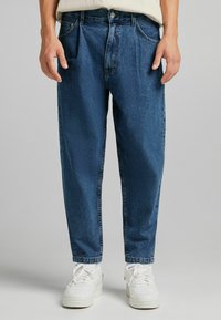 Blue denim jeans with a relaxed fit, featuring front pockets, belt loops, and a button closure; texture appears slightly faded. White sneakers below.