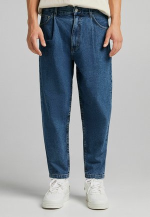 Vaqueros de denim azul con un corte relajado, que presentan bolsillos frontales, lazos para cinturón y un cierre de botón; la textura parece ligeramente desvaída. Zapatillas blancas abajo.