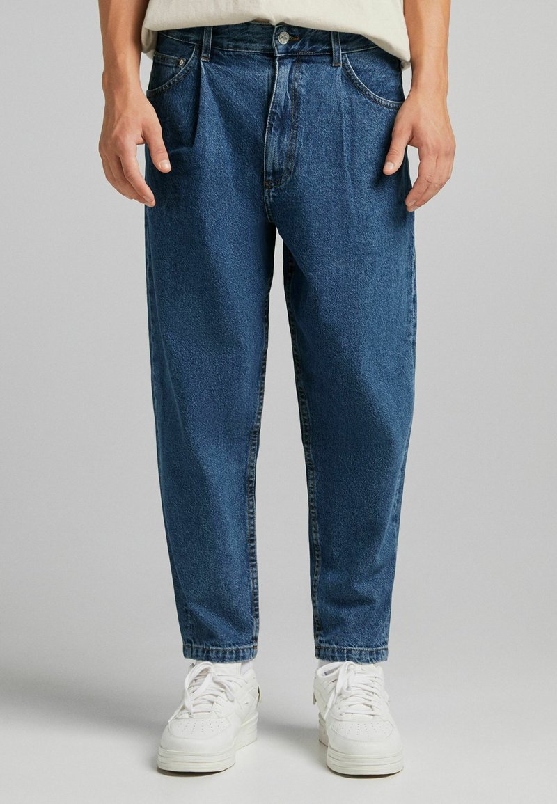 Blue denim jeans with a relaxed fit, featuring front pockets, belt loops, and a button closure; texture appears slightly faded. White sneakers below.