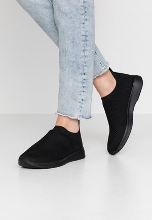 Person iført lyseblå jeans med frynset kant og sorte slip-on strikkede sneakers, stående på en glat grå gulv mod en ensfarvet baggrund.
