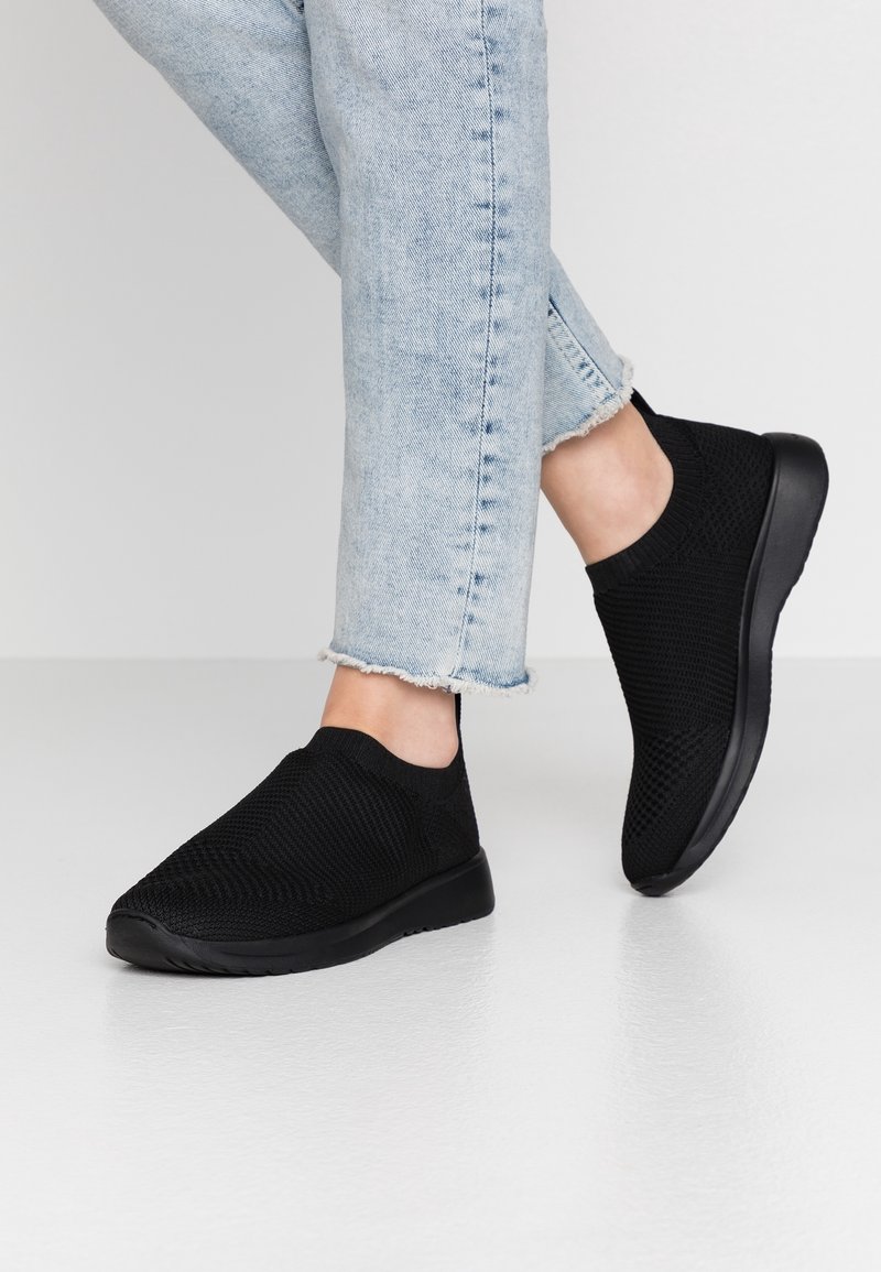Personne portant un jean bleu clair à ourlet effiloché et des baskets slip-on en maille noire, debout sur un sol gris lisse devant un arrière-plan uni.