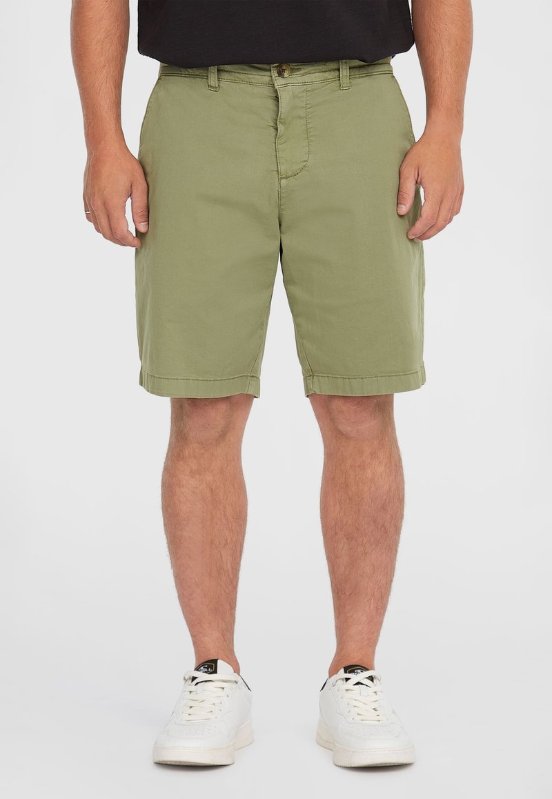 Olivgrüne Baumwollshorts mit flachem Bund, aufgerolltem Saum und seitlichen Taschen. Getragen mit weißen Sneakern, kombiniert mit einem schwarzen Shirt.