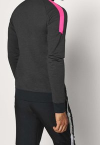 Mann in einem dunkelgrauen Sweatshirt mit einem leuchtend pinken Streifen auf der Schulter und schwarzen Hosen mit weißen Seitenstreifen, steht seitlich.