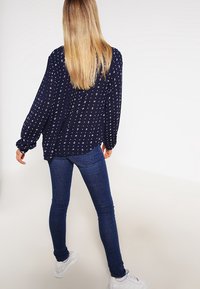 Marinebluse med hvidt diamantmønster, lange ærmer og afslappet pasform, parret med mørkeblå skinny jeans og hvide sneakers.
