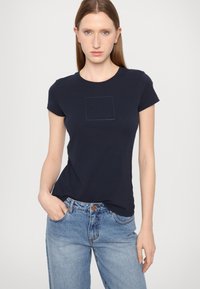 T-shirt di cotone blu navy con design aderente, maniche corte e logo testurizzato all'interno di un quadrato sul davanti. Stile casual.