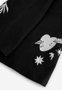 Fekete szövet fehér hímzett mintákkal, amelyek között szerepel egy levéllel díszített szív, csillagok és madarak, sima felületet és részletes varrást bemutatva.