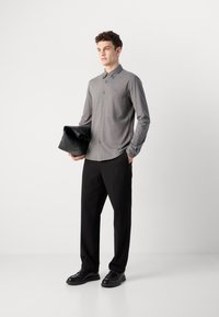 Grijze button-up overhemd met lange mouwen, gecombineerd met zwarte broeken en zwarte schoenen. Houdt een zwarte tas vast. Eenvoudig en modern ontwerp.
