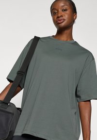 Tmavo zelené oversize tričko s okrúhlym výstrihom a krátkymi rukávmi. Má jemné logo v strede a hladkú textúru tkaniny.