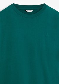 Camiseta de algodón verde azulado con cuello redondo y mangas cortas. Presenta un pequeño logo de calavera y huesos cruzados en el lado izquierdo del pecho. Textura suave.