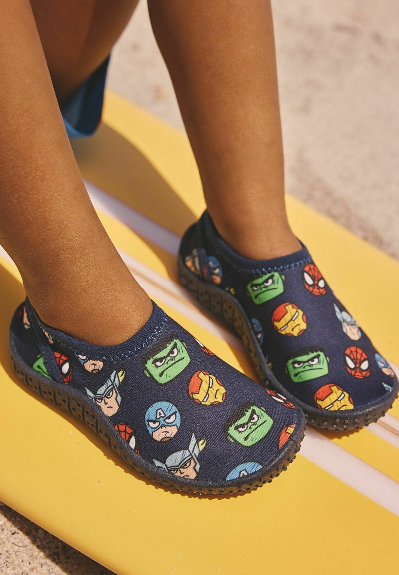 Kinderfüße in marineblauen Slip-On-Schuhen mit Marvel-Superheldengesichtern, stehend auf einem gelben Surfbrett auf sandigem Boden.