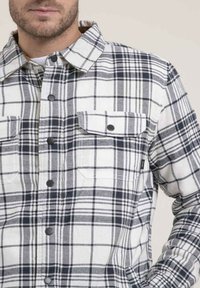 Chemise en flanelle à carreaux blanche et marine. Comprend deux poches poitrine, un col et une fermeture à boutons sur le devant. Tissu texturé.