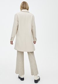 Cappotto in misto lana beige con taglio dritto, colletto a rever e polsini con bottone. Presenta una texture morbida e arriva sotto i fianchi.