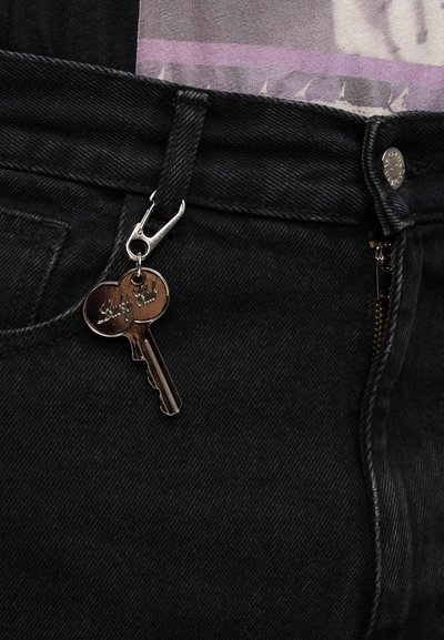 Μαύρο τζιν παντελόνι με ασημένιο φερμουάρ, που έχει κλιπ μπρελόκ με μεταλλικό σχέδιο κλειδιού. Το ύφασμα παρουσιάζει υφή.