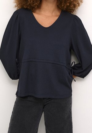 Femme portant un chemisier à manches longues bleu marine foncé avec des manches bouffantes et des détails de lien sur le côté, associé à un jean noir délavé.