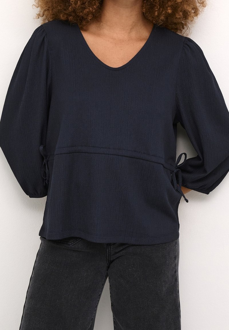 Femme portant un chemisier à manches longues bleu marine foncé avec des manches bouffantes et des détails de lien sur le côté, associé à un jean noir délavé.