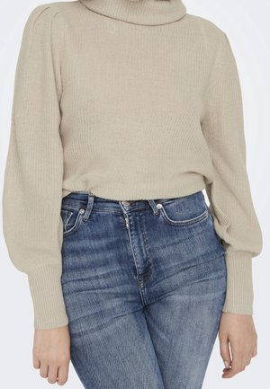 Beige gebreide coltrui met opgeblazen lange mouwen, gecombineerd met hoogwaterverlopen blauwe jeans bij een persoon.