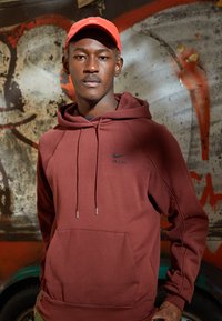 Vinröd Nike-hoodie med känguruficka, dragsnöre och logotyp på bröstet. Klargul keps kompletterar outfiten. Texturerad tyg.