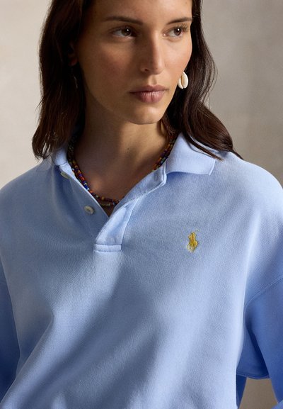 Mujer con camisa polo azul claro con logo bordado amarillo, pendiente de concha y collar colorido de cuentas, mirando hacia su izquierda.
