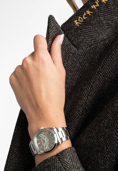 Srebrny zegarek ze stali nierdzewnej z ciemnoszarym cyferblatem z motywem skrzydeł, noszony na ręce obok teksturowanej ciemnej marynarki z wełny.