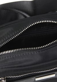 Svart tygväska med synliga dragkedjor och slät foder; har silverdetaljer, flera fack och en märkesetikett.