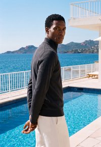 Homme en pull sombre et pantalon blanc debout près d’une piscine avec l'océan et des montagnes en arrière-plan, regardant par-dessus son épaule.