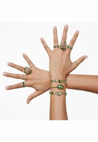 Multiple hands adorned with various rings and bracelets. Materials include gold and gemstones, featuring green and pink colors and intricate designs.