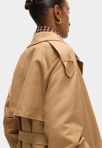Beige Trenchcoat mit einem zweireihigen Design, großen Klappen und einem taillierten Gürtel, aus glattem Stoff und mit einem karierten Kragen.