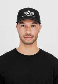 Fekete snapback sapka ívelt szélekkel, rajta az "ALPHA INDUSTRIES" fehér logójával és vonalakkal. Fekete kerek nyakú pólóval viselve.