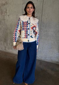 Γυναίκα που στέκεται μέσα σε κλειστό χώρο, φοράει πουλόβερ με κουμπιά και σχέδιο, με γιακά από shearling, φαρδιά σκουρόχρωμα τζιν και κρατά μια μπεζ τσάντα.