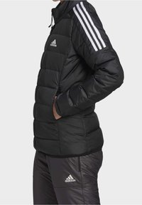 Černá prošívaná bunda s bílým třemi pruhy na ramenou, vybavená zipem a přední kapsou. Hladká, prošívaná textura.