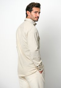 Lichtbeige langdofshirt met een kraag, gestructureerde stof en knoopaccenten op de mouwen, van achteren getoond, ingestopt in lichte broeken.