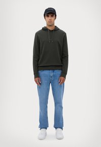 Mørkegrønn hettegenser med snor, lyseblå jeans og hvite sneakers. Modell iført en mørk caps, stående mot en nøytral bakgrunn.