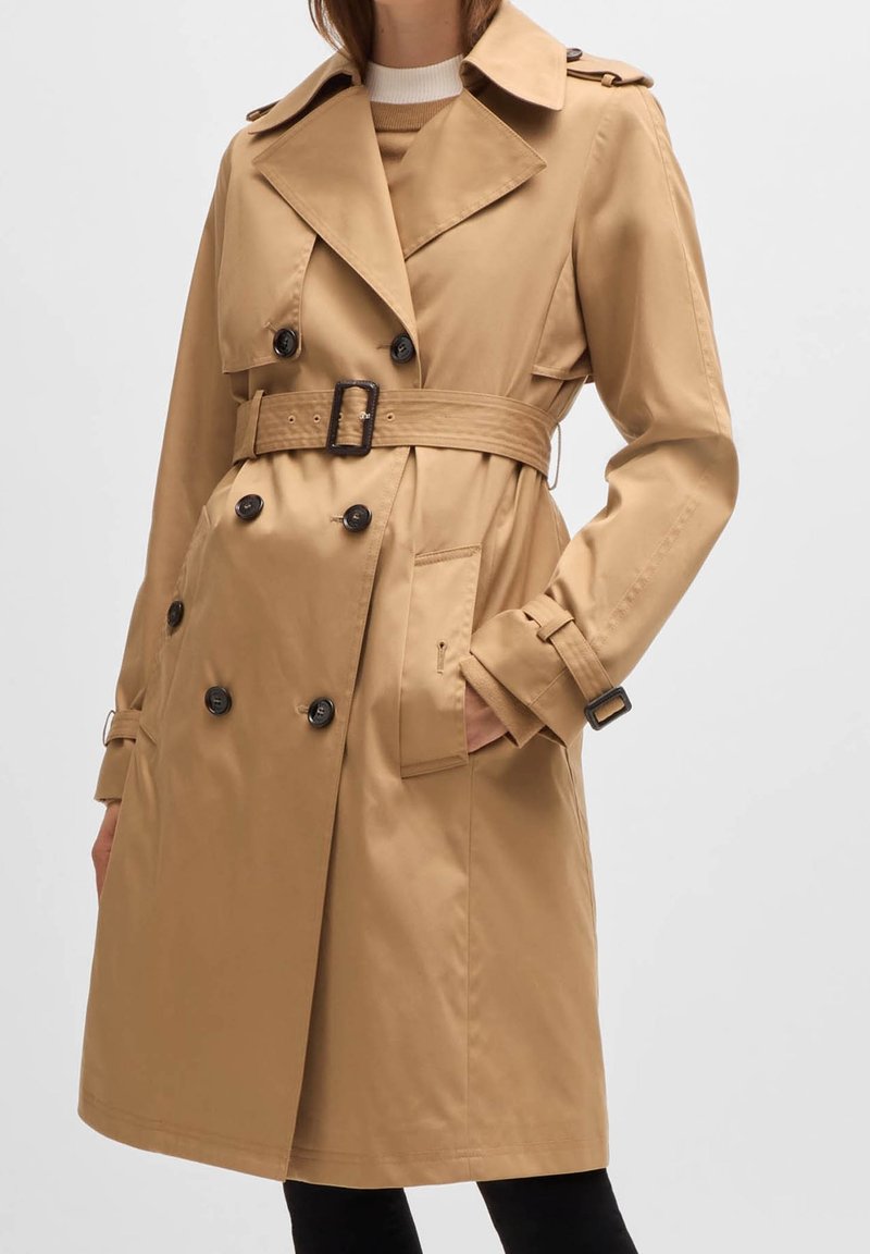Femme portant un trench-coat beige à double boutonnage avec ceinture, boutons noirs et sangles aux poignets, main dans la poche sur fond blanc.