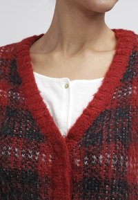 Cárdigan de punto con patrón rojo y negro, de escote en V profundo, con una textura suave y detalles de botones visibles sobre una blusa blanca.