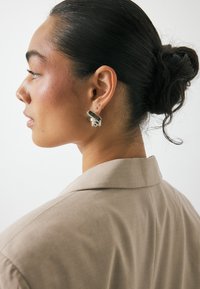 Ασημένιες σκουλαρίκια κρίκοι με σπειροειδή σχέδιο και έντονες ραβδώσεις. Το μοντέλο φοράει ένα ανοιχτό μπεζ σακάκι με διαρθρωμένο γιακά.