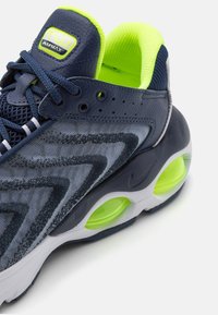 Marinblå sportskor med överdel i mesh och tyg, med en kurvad design, gröna detaljer, synliga luftpoddar för dämpning och texturerad utsmyckning.