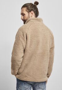Kreemivärvi fliistekott, millel on tekstureeritud, karvane pind, kõrge krae ja lõdva lõikega. Kandmisel sinised teksad.
